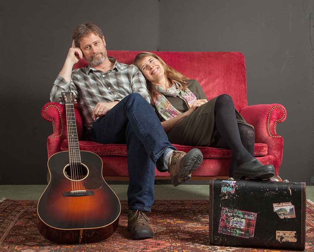 Musicians Dana and Susan Robinson on a red couch with a guitar and case at their feet.